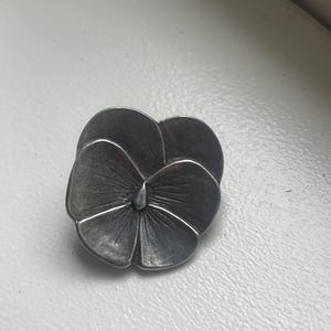 May include: A detailed, silver-toned metal flower brooch. The brooch is shaped like a pansy, with detailed petal lines and a central bud. The metal has a slightly aged appearance, giving it a vintage feel. The brooch is a decorative accessory.