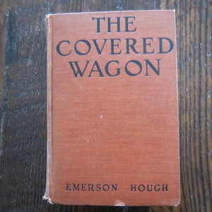 Puede incluir: Un libro vintage titulado "The Covered Wagon" de Emerson Hough. El libro tiene una cubierta roja con texto negro.