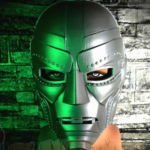 May include: A silver-colored metallic mask featuring a rigid, segmented design with prominent brow plates, a central nose bridge, and a horizontal mouth opening. The mask displays rows of small circular rivets along the eye and jaw sections.
