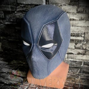 May include: A full-head mask featuring a textured dark grey finish with smooth, glossy black eye panels and white mesh eye inserts. The mask is displayed on a tan-coloured stand, showcasing a contoured design with defined seams and panels.