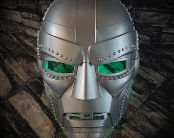 May include: A metallic silver full-face mask featuring a rigid, segmented design with prominent rivet-like detailing along the brow and cheek areas. The mask includes rectangular eye openings with bright green lenses and a horizontal mouth vent.