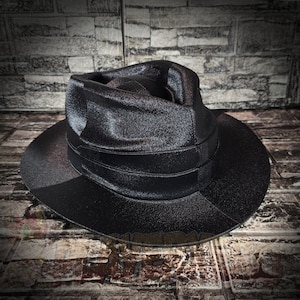 Puede incluir: Un sombrero fedora negro con un acabado brillante de aspecto metálico. El sombrero presenta una corona pellizcada y un ala ancha y estructurada. Una banda negra a juego envuelve la base de la corona, aportando un aspecto uniforme.