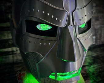 Puede incluir: Un casco integral de color plata metálica con un diseño rígido y segmentado, que presenta detalles de remaches prominentes a lo largo de la frente y las mejillas. Las cuencas de los ojos y la boca están iluminadas con una luz verde brillante.