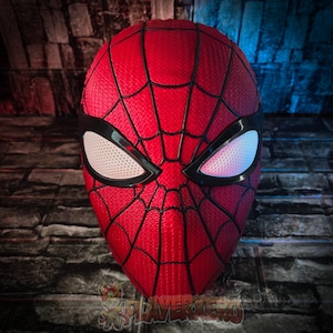 Könnte beinhalten: Eine rote Spider-Man-Maske mit schwarzem Spinnennetz-Muster und schwarzen Augenrändern. Die Augenlinsen sind weiß mit einer gepunkteten Textur. Die Maske wird vor einem Backsteinmauer-Hintergrund gezeigt.