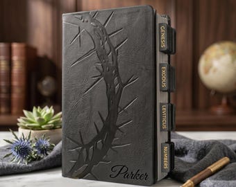 May include: A black leather-bound book featuring an embossed crown of thorns design on the front cover and the name Parker engraved at the bottom. The book includes black index tabs labeled Genesis, Exodus, Leviticus, and Numbers in gold lettering.