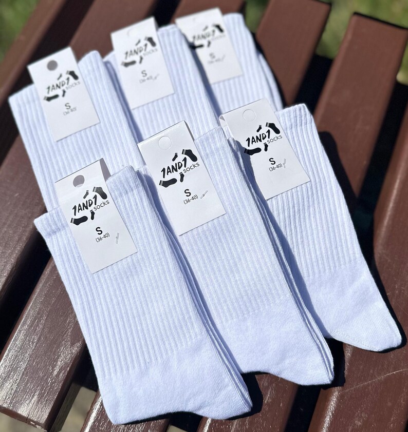 May include: A set of five pairs of white ribbed crew socks, each featuring a white cardboard tag with the black text 1AND1 socks and the size S (36-40) printed on the front. The socks are arranged in a layered pattern, showcasing the fabric texture.