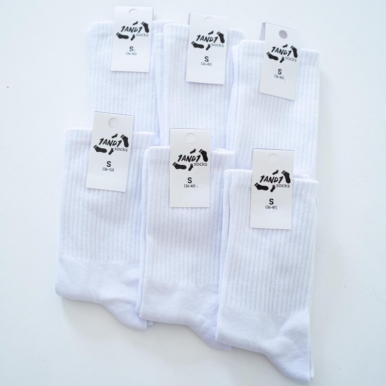 May include: Six pairs of solid white ribbed crew socks arranged in a group. Each pair features a white paper tag attached to the top with the black text 1AND1 socks, the letter S, and the numerical range 36-40 printed clearly on the front.