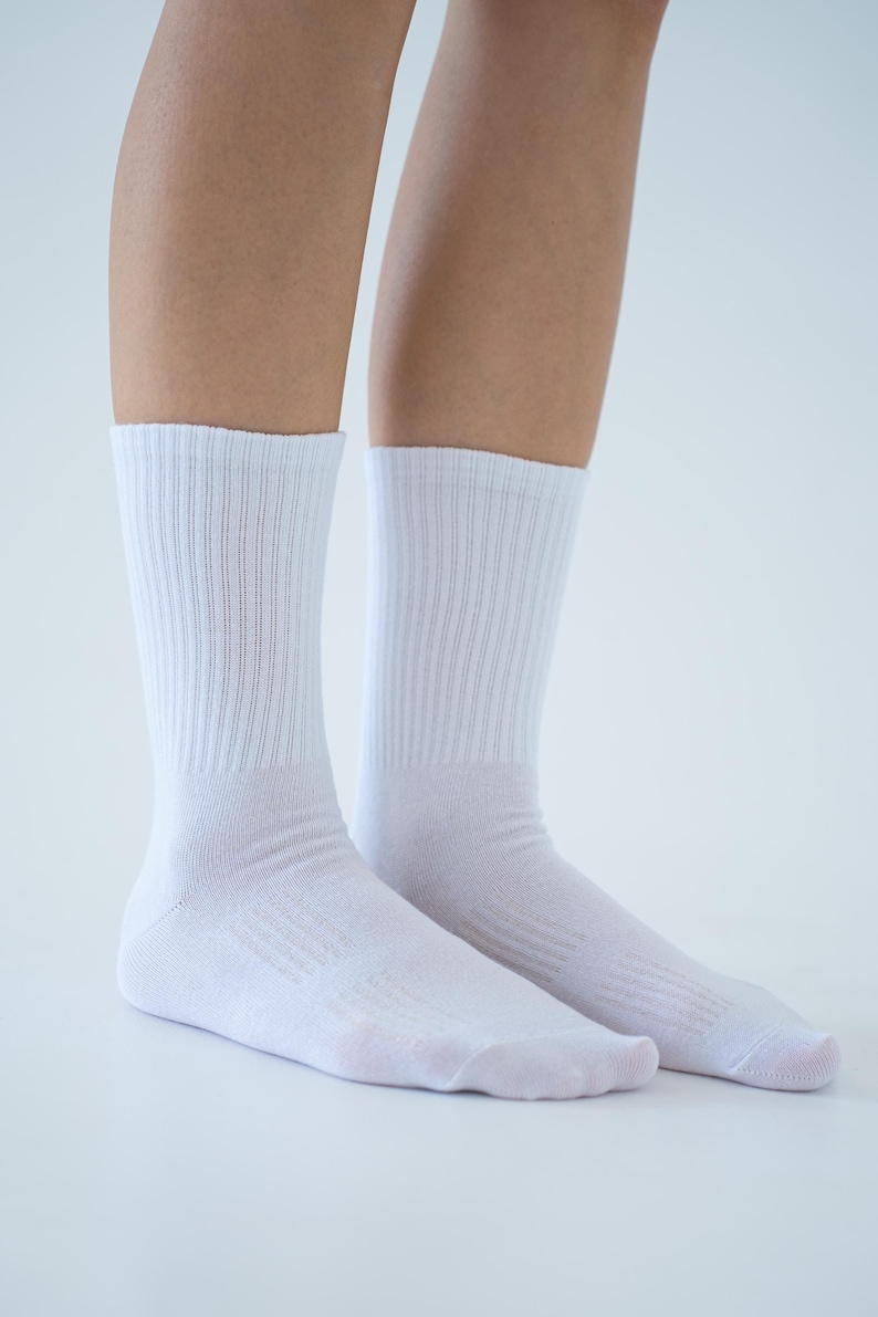 May include: A pair of solid white crew socks featuring a ribbed knit texture on the upper leg portion and a smooth, plain knit finish on the foot area. The socks extend to the mid-calf and are constructed from a lightweight, opaque fabric.