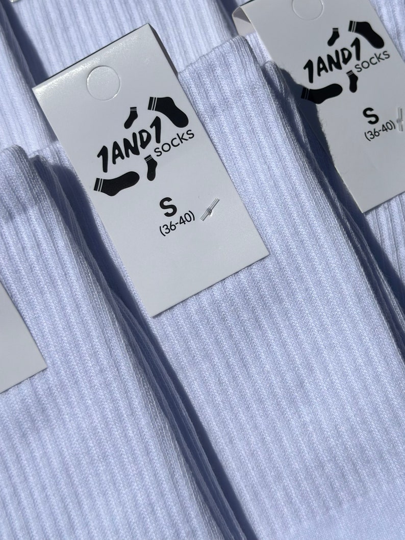 May include: A pair of white ribbed socks with a white paper tag attached. The tag features the black text 1AND1 socks, a graphic of a sock, and the size S (36-40) printed clearly on the front. The fabric has a vertical ribbed texture.