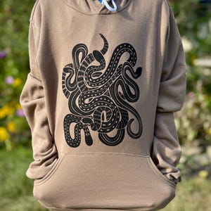 Peut inclure: Un sweat à capuche beige avec un motif de poulpe noir. Le sweat à capuche a une capuche à cordon, une poche kangourou et des manches longues. Le motif de poulpe est une illustration stylisée et détaillée.