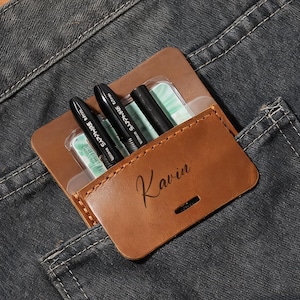 May include: A brown leather pen holder with the name "Kavin" in cursive is in the pocket of a pair of gray jeans. The pen holder contains several black pens and green and white cards. The background is a wooden surface.