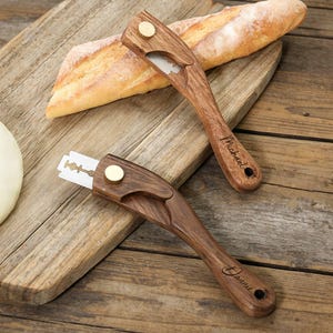 May include: Two wooden bread lame tools with exposed blades and brass accents rest on a wooden cutting board with a loaf of bread. One tool is engraved with the name "Michael", the other with "Dannie". A loaf of bread and a baguette are also on the board.