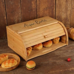 May include: A bamboo bread box with a roll-top door, holding various baked goods. The box has the name "Alison Bartel" engraved on the lid. A wooden plate with bread and a small burger are in the foreground.