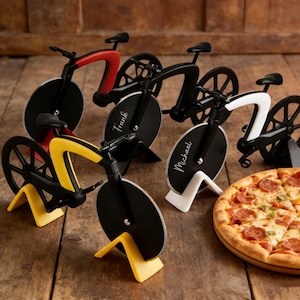 Könnte beinhalten: Eine Auswahl an Fahrrad-förmigen Pizzaschneidern in Rot, Gelb, Schwarz und Weiß. Jeder Schneider hat eine kreisförmige Klinge und steht auf einer Holzoberfläche. Eine Pizza ist ebenfalls zu sehen.