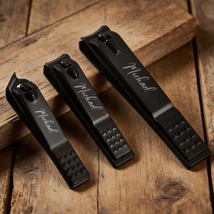 May include: Three black nail clippers of varying sizes, each engraved with the name "Michael" in a cursive font. The clippers are arranged on a rustic wooden surface, showcasing their sleek, matte finish and textured grip areas.