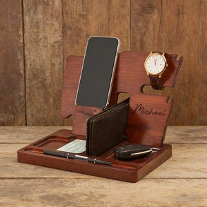 Könnte beinhalten: Ein Schreibtisch-Organizer aus dunkelbraunem Holz. Er hat Fächer für ein Smartphone, eine Geldbörse und eine Uhr sowie eine Ablage für kleine Gegenstände. Eine goldfarbene Uhr mit braunem Lederarmband liegt seitlich. Der Name "Michael" ist eingraviert.