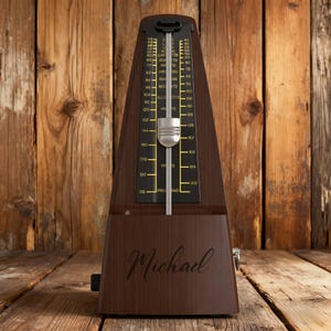 Könnte beinhalten: Dunkelbraunes Holzmetronom mit schwarzem Zifferblatt und silbernem Pendel. Das Zifferblatt zeigt Tempoangaben und der Name "Michael" ist auf der Vorderseite eingraviert. Das Metronom steht vor einem Holzuntergrund.