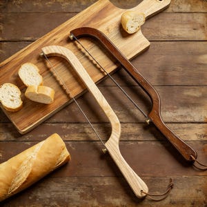 Peut inclure: Deux scies à pain en bois avec des fils en acier inoxydable, une claire et une foncée, reposent sur une planche à découper en bois avec des tranches de pain. Une baguette est également présente, suggérant une cuisine ou une salle à manger.