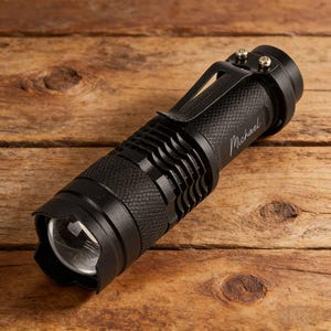 May include: Black flashlight with a textured grip and a metal clip. The name "Michael" is inscribed on the side. The flashlight is lying on a wooden surface.