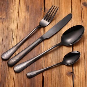 May include: A set of four black stainless steel utensils: a fork, a knife, a large spoon, and a small spoon. Each piece is engraved with a name in a cursive font. The utensils are arranged on a wooden surface.