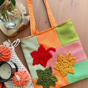 Può includere: Una borsa tote patchwork con manici arancioni e un design colorato. La borsa presenta quadrati di tessuto verde, arancione e rosa, adornati con applicazioni di foglie all'uncinetto rosse, gialle e verdi. Un articolo stagionale fatto a mano.