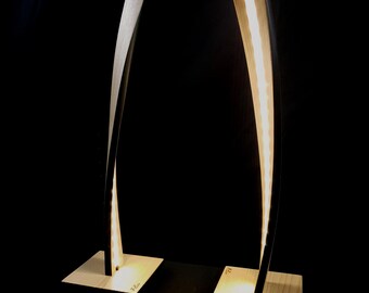 May include: Two modern wooden lamps with a curved design. The lamps are lit and have a warm white glow. The bases are made of light wood and the lamps are on a black surface.