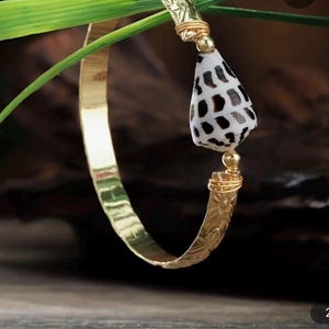 May include: A gold-coloured bangle bracelet with a seashell pendant. The bracelet has a hammered texture and a decorative design near the pendant. The pendant is a white seashell with black spots. The bracelet is displayed against a blurred background.