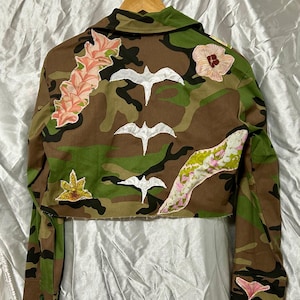 Pode incluir: Casaco camuflado com aplicações florais e de pássaros. O casaco apresenta um padrão de camuflagem clássico em tons de verde, castanho e bege. As costas são adornadas com desenhos de pássaros brancos e patches florais rosa e amarelos. As mangas são compridas e o casaco é curto.