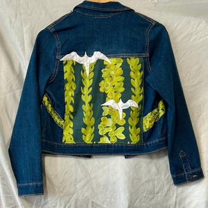 May include: A dark blue denim jacket with a decorative back panel featuring a green floral design and white bird appliqués. The jacket has a classic collar, button closures, and pockets.