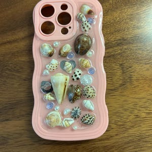 May include: A pink phone case decorated with various seashells, pearls, and small blue beads. The case has a wavy edge and cutouts for the phone's camera and buttons. The seashells are in different shapes, sizes, and colors.