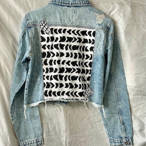 May include: Light blue denim jacket with a distressed design. The back features a white panel with a black abstract pattern. Two small black and white patches are on the jacket. The jacket has a raw hem and a button closure.