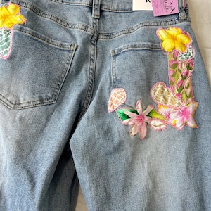 May include: Light blue denim jeans with floral embroidery on the back pockets. The embroidery features yellow, pink, and green flowers with intricate details. The jeans have a classic five-pocket design and a button and zipper closure.