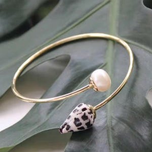 May include: A gold-toned bracelet featuring a white pearl and a seashell with black and white markings. The bracelet has a delicate, open design, resting on a green leaf.