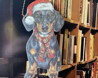 May include: A black and white dachshund wearing a red and white Santa hat and a red and white scarf. The dachshund is a Christmas ornament.