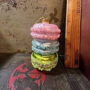 May include: A decorative ornament shaped like a stack of macarons. The ornament features layers of pink, blue, and green, each with a ruffled edge. A gold-colored loop is at the top for hanging.