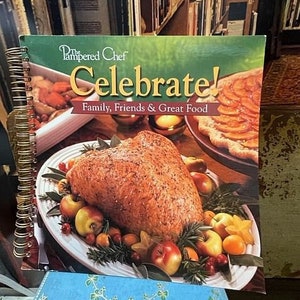 May include: A cookbook titled "The Pampered Chef Celebrate! Family, Friends & Great Food" with a spiral binding. The cover features a roasted turkey breast with apples, cranberries, and rosemary on a white platter.