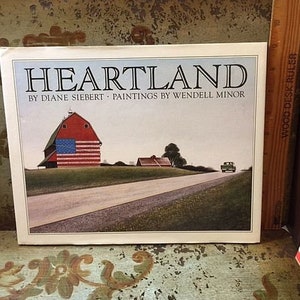 May include: A book cover with the title "HEARTLAND" by Diane Siebert - Paintings by Wendell Minor. The cover features a red barn with an American flag painted on it, a country road, and a green truck.