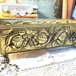 May include: A rectangular, gold-colored metal planter with embossed rose designs. The planter sits on four small, claw-shaped feet. The sides and front of the planter feature detailed floral relief patterns, creating a vintage aesthetic.