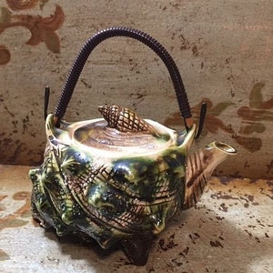 May include: A ceramic teapot shaped like a seashell with a brown handle and a green and brown glaze. The teapot has a small seashell on the lid.