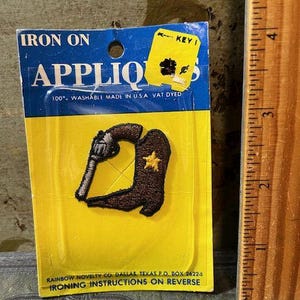 May include: An iron-on applique featuring a brown cowboy boot with a yellow star and a silver revolver. The applique is on a yellow and blue card with text that reads "Iron On Appliques". The card also includes the text "100% Washable Made in USA".