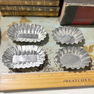 May include: Four vintage, silver-colored, fluted metal baking molds. The molds are rectangular with a scalloped edge. A wooden ruler with the word "WESTCOTT" is in the foreground, and several antique books are in the background.