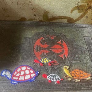 May include: Four embroidered turtle patches in various colors, including blue, red, yellow, and orange, are displayed on a dark, textured surface. The patches are arranged near a circular design with a red background and black figures.