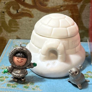 May include: A white igloo-shaped ornament with a small opening, accompanied by miniature figurines of an Inuit person and a seal. The Inuit figure is wearing a hooded parka and mittens. The seal is gray. The igloo has the number "2009" on it. The items are on a blue surface.