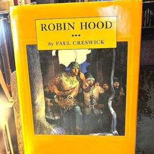May include: A hardcover book titled "ROBIN HOOD" by Paul Creswick. The cover is a vibrant yellow with a framed illustration depicting Robin Hood and his men in a forest setting. The book is resting on a white lace tablecloth.