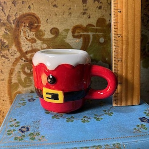 May include: A small, red ceramic mug with a white rim and a Santa Claus design. The mug features a black belt with a yellow buckle and a black eye. The mug is on a blue book.