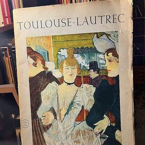 May include: A vintage book cover featuring the name "TOULOUSE-LAUTREC" in large, bold letters. The cover displays a colorful illustration of several figures in a cafe setting, with a woman in a white dress as the focal point. The book is aged with visible wear.