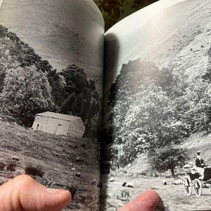 May include: Black and white photograph of a landscape scene. A small stone building sits on a hillside, surrounded by trees. A horse-drawn carriage with two horses and a driver is in the foreground.