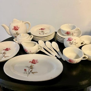 May include: A vintage tea set with a floral rose design. The set includes a teapot, sugar bowl, creamer, plates, cups, saucers, and spoons. The dishes are white with a red rose and green stem design.