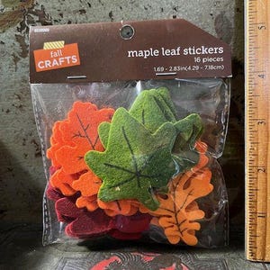 May include: A clear plastic bag contains 16 felt maple leaf stickers in shades of orange, green, and burgundy. The package reads "maple leaf stickers" and "fall crafts". The leaves measure 1.69 - 2.83 inches.