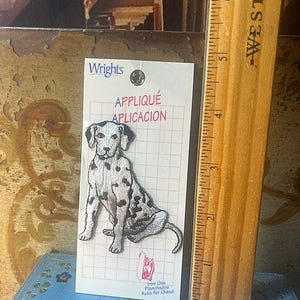 May include: An iron-on applique patch featuring a Dalmatian dog with black spots, sitting on a white background with a grid pattern. The patch is labeled "Wrights" and includes instructions in English, Spanish, and French. A wooden ruler is next to the patch.